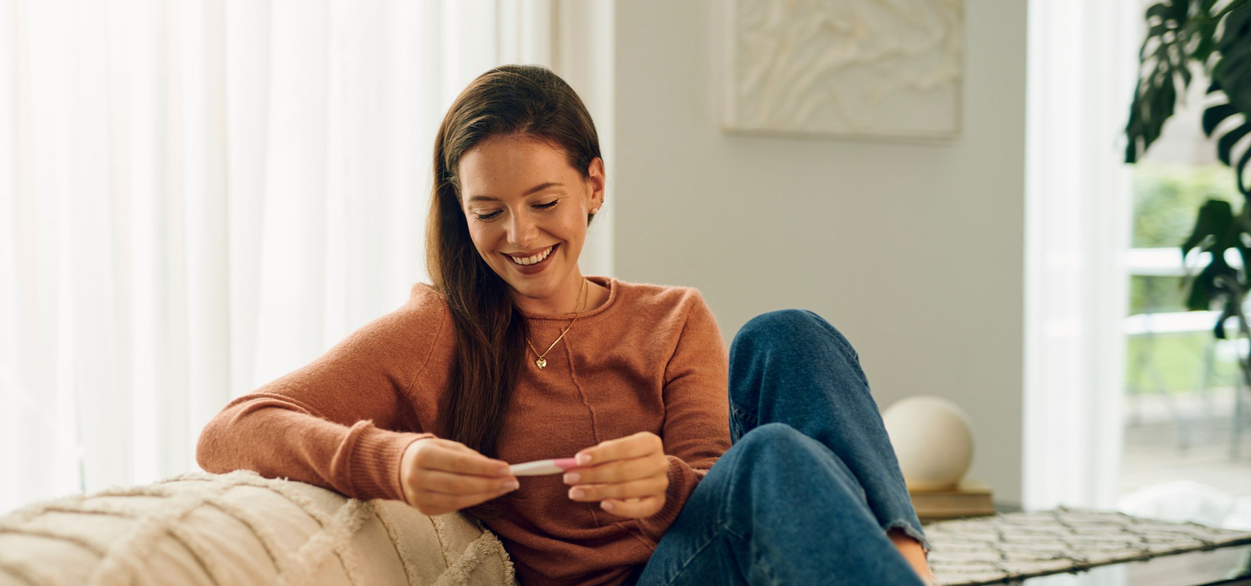 Бременна жена, седнала на дивана, гледайки положителен тест за бременност, докато планира бременност