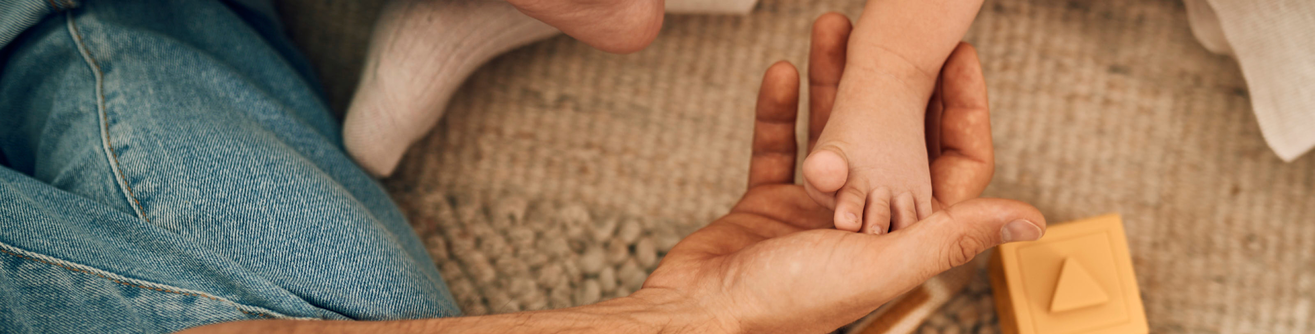 Татко държи крачето на бебето, мек килим и играчки