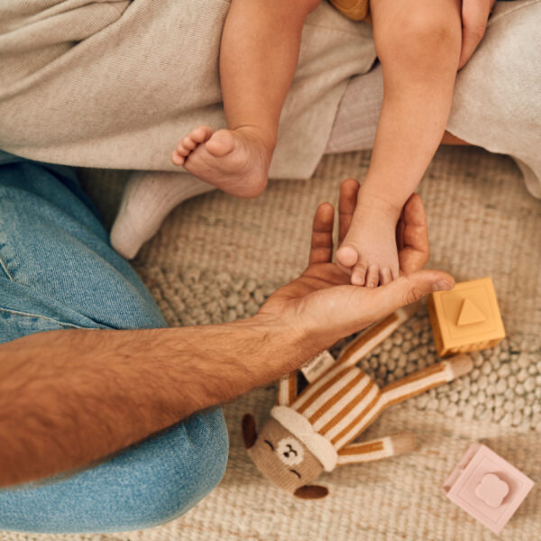 Татко държи крачето на бебето, мек килим и играчки