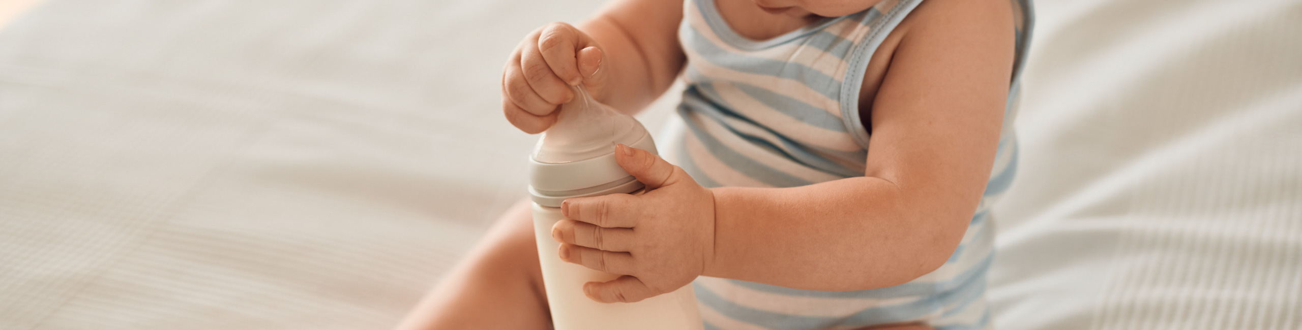 Бебето седи на леглото и държи шише в ръце.