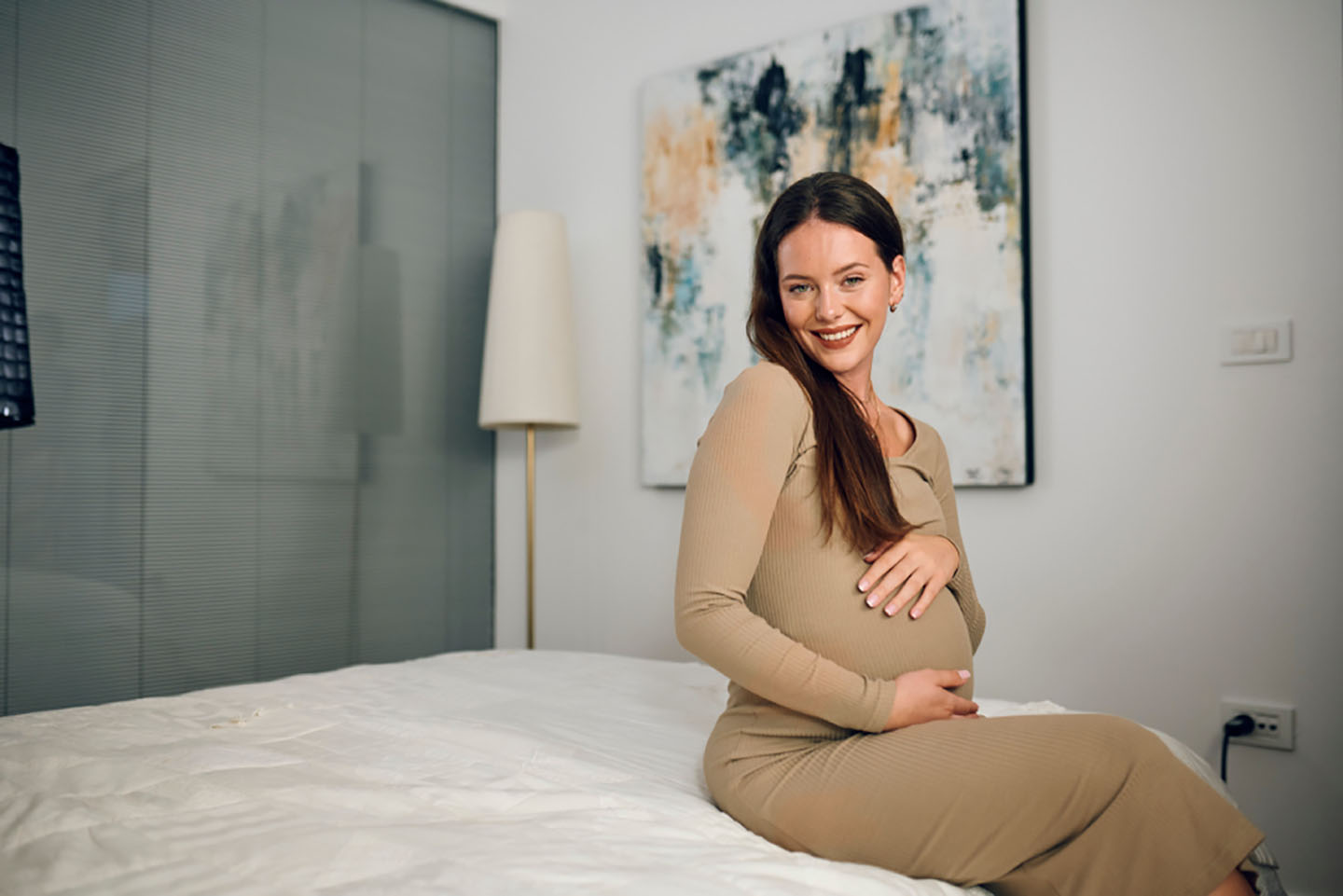 Бременна жена седи на легло и се усмихва