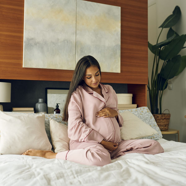 Бременна жена в розов халат седи на легло и държи корема си.